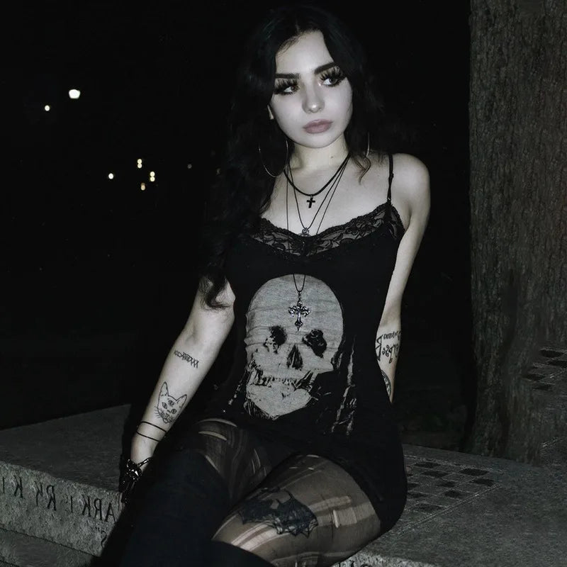 Black skull print lace mini dress worn by a seated model at night. The pale skull, delicate neckline and patterned tights stand out against the dark background.