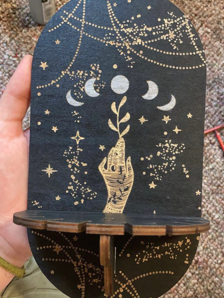 Small black wooden shelf held in one hand, showing an illustrated hand beneath moon phases, gold-tone stars and a notched projecting ledge.