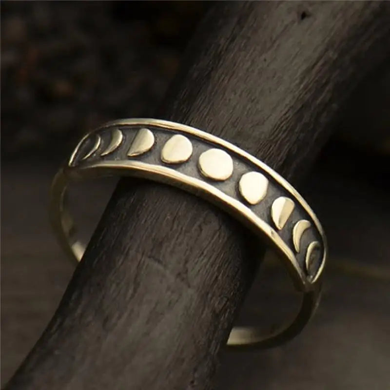 Moon phase band ring resting around a dark wooden branch. Raised lunar shapes catch warm light above a dark recessed strip.