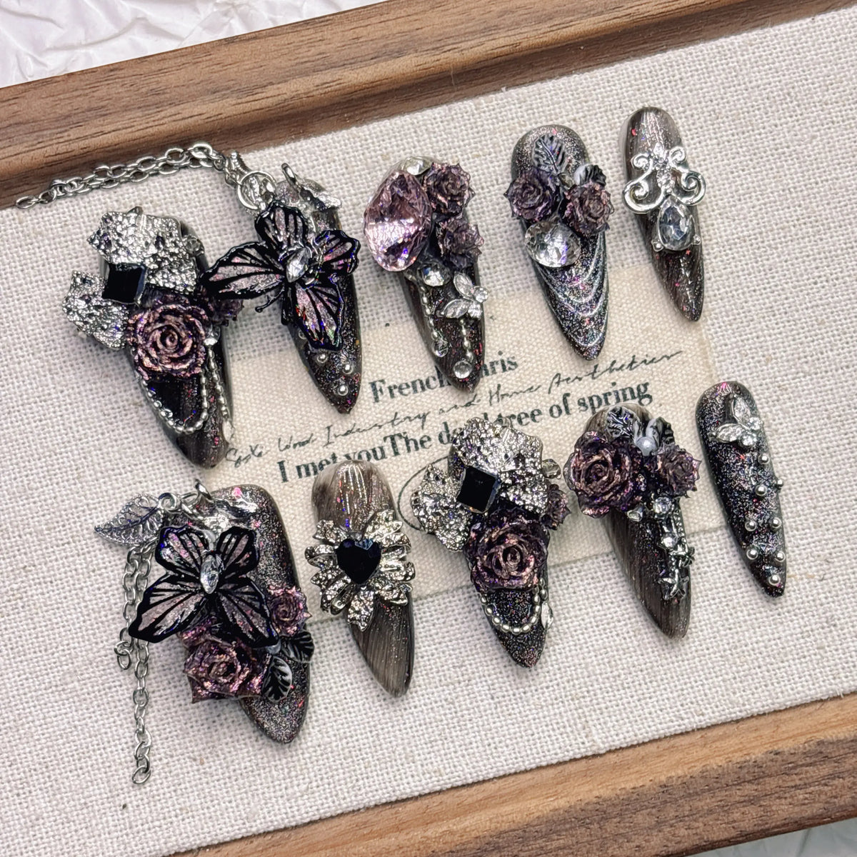 Purple and black almond press-on nails arranged on a pale fabric-lined wooden tray, with raised roses, butterfly wings, faceted accents and fine silver-tone chains.