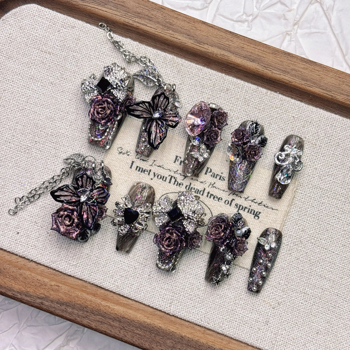 Gothic stiletto press-on nails displayed at an angle on a wooden tray, showing pointed black-purple tips, sculpted roses, butterfly accents and hanging chains.
