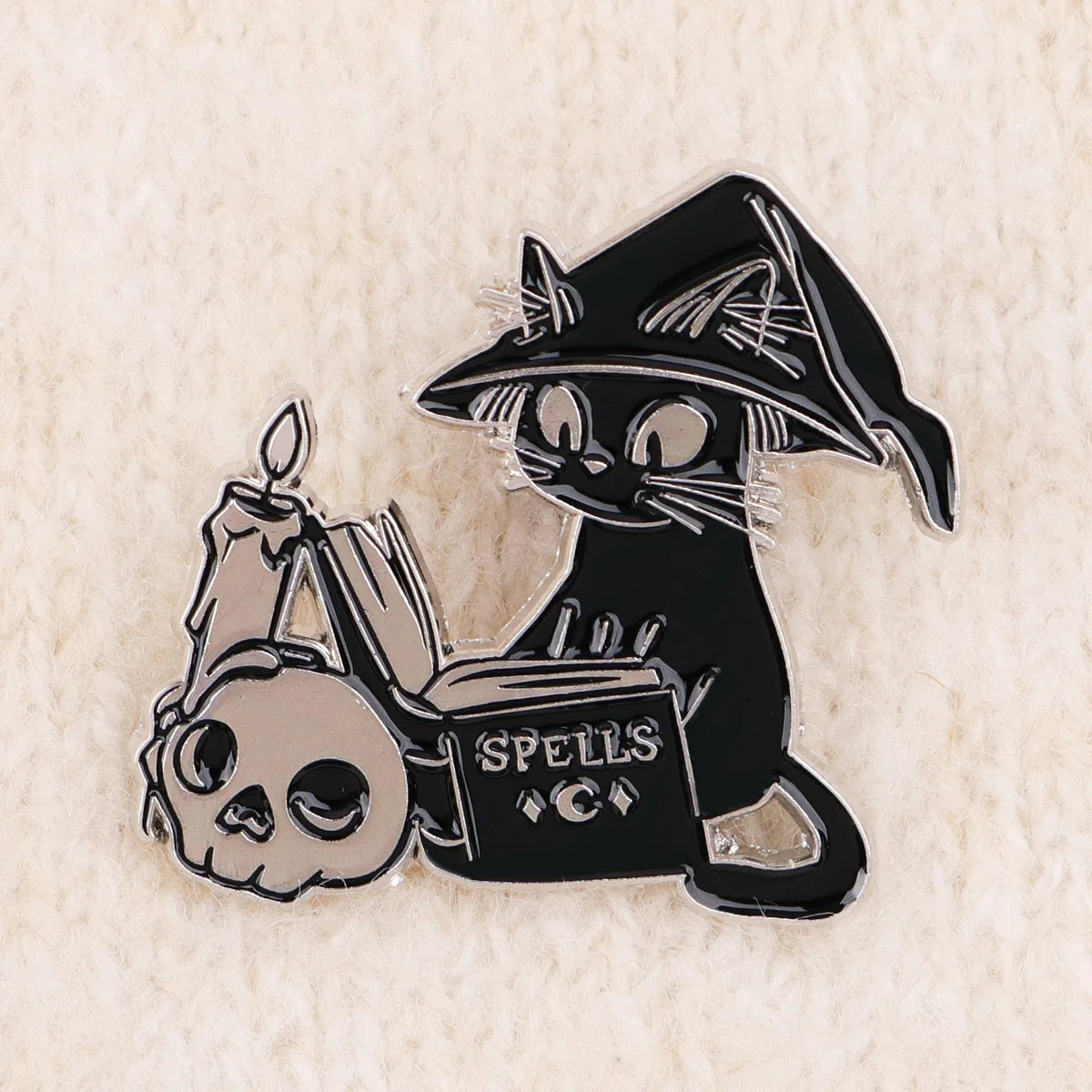 Black witch cat enamel pin on a textured cream background, with spellbook and candle details