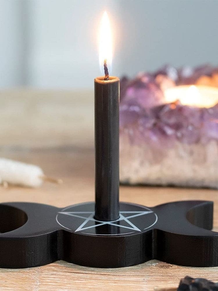 Black triple moon candle holder with a lit dark candle in the central opening, shown on wood with a pale pentagram around the base.