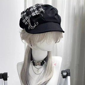 Black gothic newsboy cap with a monochrome plaid patch, safety-pin accents, draped chains and a curved brim.