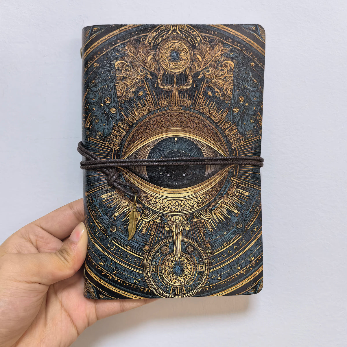 Front view of the all-seeing eye notebook against a pale background, showing the dark iris and elaborate gold-coloured linework.