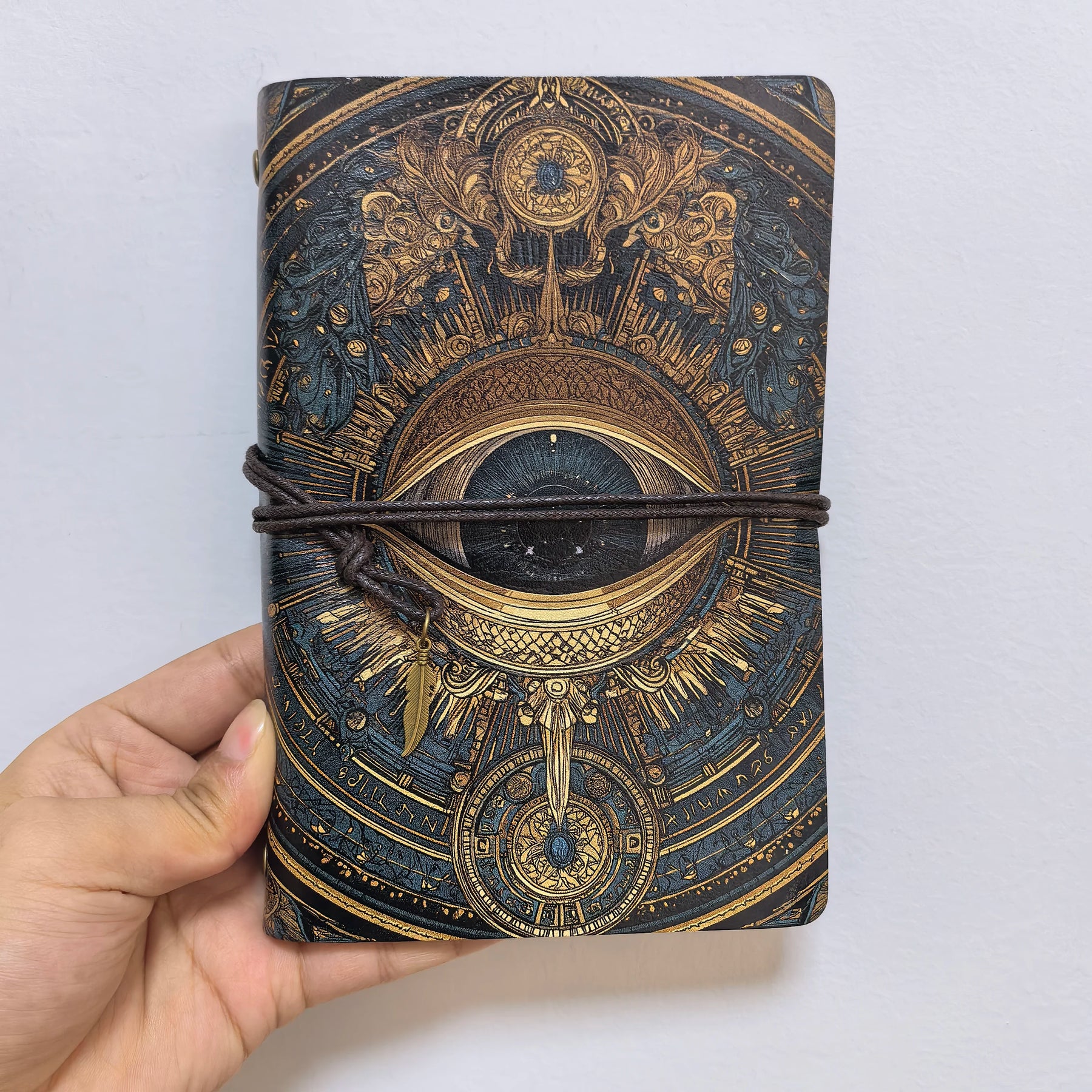 Front view of the all-seeing eye notebook against a pale background, showing the dark iris and elaborate gold-coloured linework.