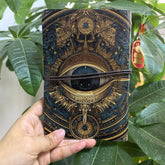 Dark blue-black refillable journal with a large eye, ornate gold-coloured borders and a cord wrapped across the cover.