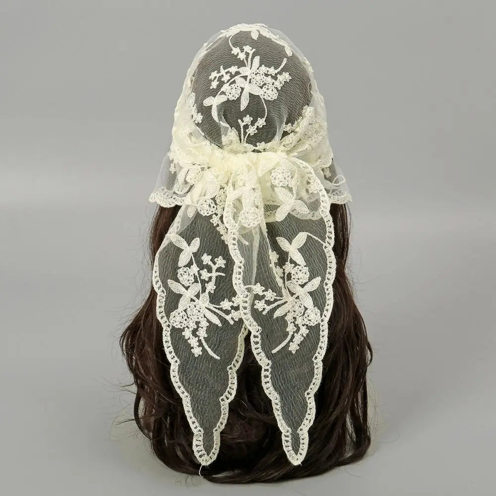 Beige floral lace headscarf tied behind dark hair against a grey background. The bow and long pointed ends show the open flower-and-vine pattern and scalloped edging.