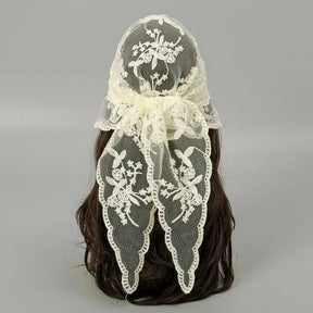 Beige floral lace headscarf tied behind dark hair against a grey background. The bow and long pointed ends show the open flower-and-vine pattern and scalloped edging.
