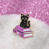 Black cat enamel pin perched on purple spellbooks, photographed on white fluffy fabric beside a pink glitter surface.