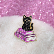 Black cat enamel pin perched on purple spellbooks, photographed on white fluffy fabric beside a pink glitter surface.
