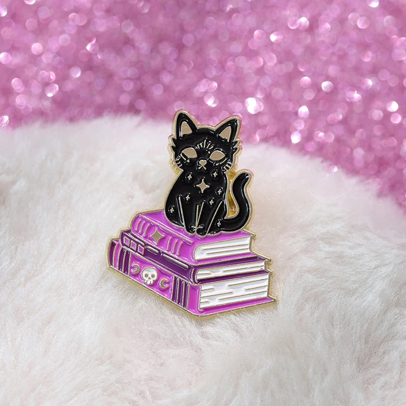 Black cat enamel pin perched on purple spellbooks, photographed on white fluffy fabric beside a pink glitter surface.