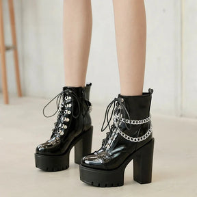 Glossy black platform ankle boots worn on a pale floor, with silver-tone ankle chains, front lacing and tall block heels clearly visible.