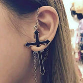 Side view of the black cross earring on an ear in warm light. A fine chain follows the upper ear and another curve hangs below the split cross.