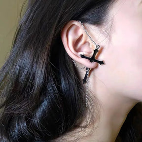 Black split cross earring worn outdoors with the fine upper chain draped over the ear. The lower cross and hanging chain stand out beside dark hair.