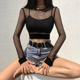 Black long sleeve fishnet crop top worn over a separate opaque black bralette with denim shorts. The seated view shows the round neckline, sheer diamond mesh and solid cuffs and hem.