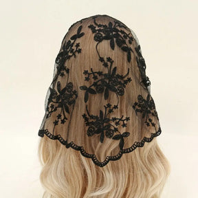 Black floral lace triangle headscarf draped over blonde hair against a pale background. The open floral pattern and scalloped border stand out clearly over the lighter hair.