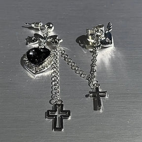 Black heart and bow earring laid beside its winged heart cuff on a gray surface. The silver-tone chains, sparkling heart border and hanging crosses are visible from above.