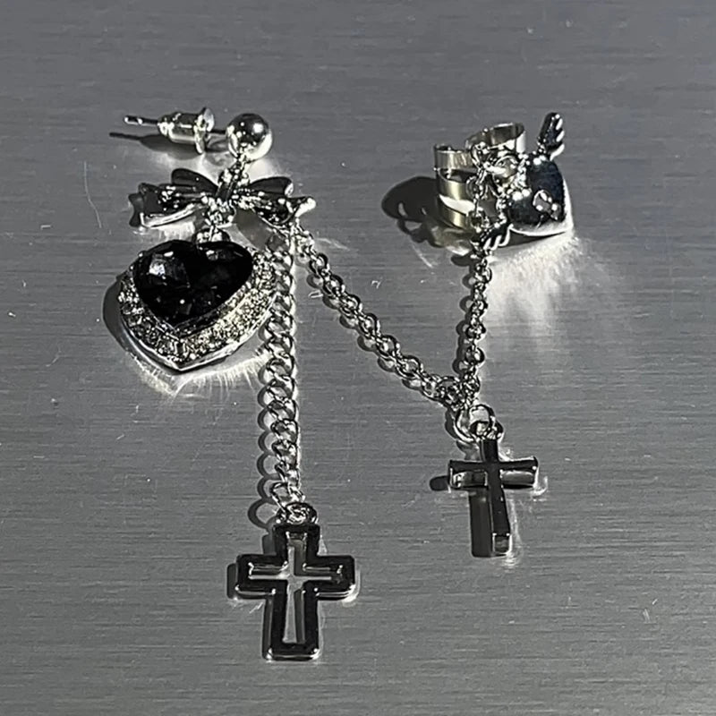 Black heart and bow earring laid beside its winged heart cuff on a gray surface. The silver-tone chains, sparkling heart border and hanging crosses are visible from above.