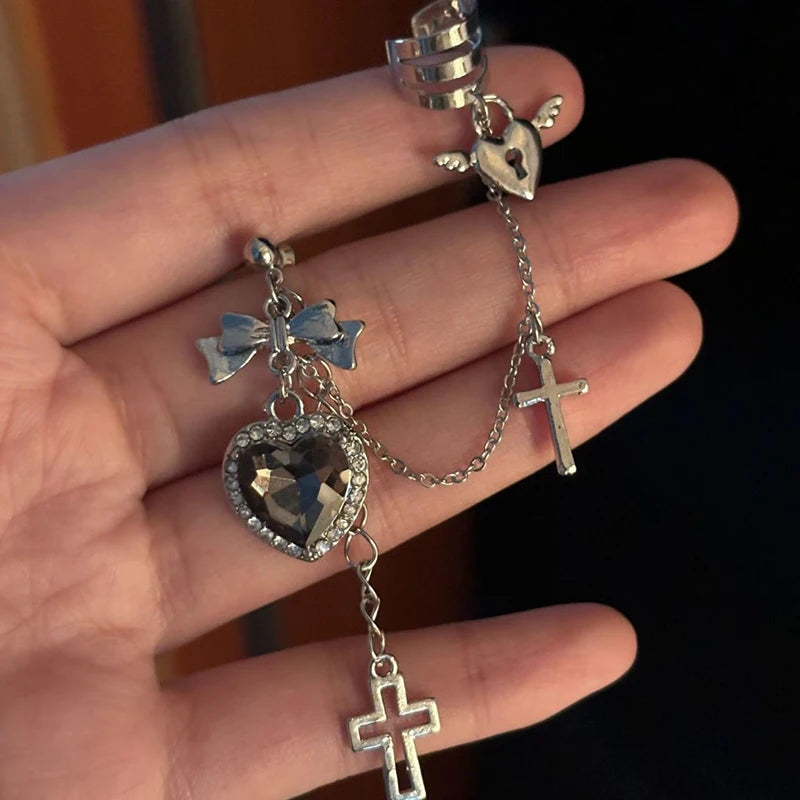 Black heart and bow earring held across a hand, with its chain extending to a winged heart ear cuff. Two different cross drops finish the silver-tone design.