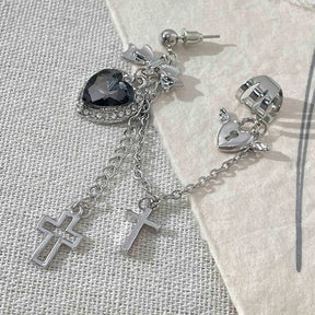 Black heart earring laid on pale textured fabric with its post and backing visible above the bow. A chain joins the winged heart cuff, with two silver-tone cross drops below.
