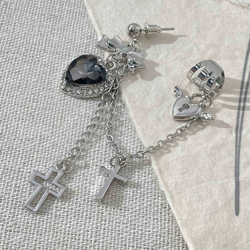 Black heart earring laid on pale textured fabric with its post and backing visible above the bow. A chain joins the winged heart cuff, with two silver-tone cross drops below.