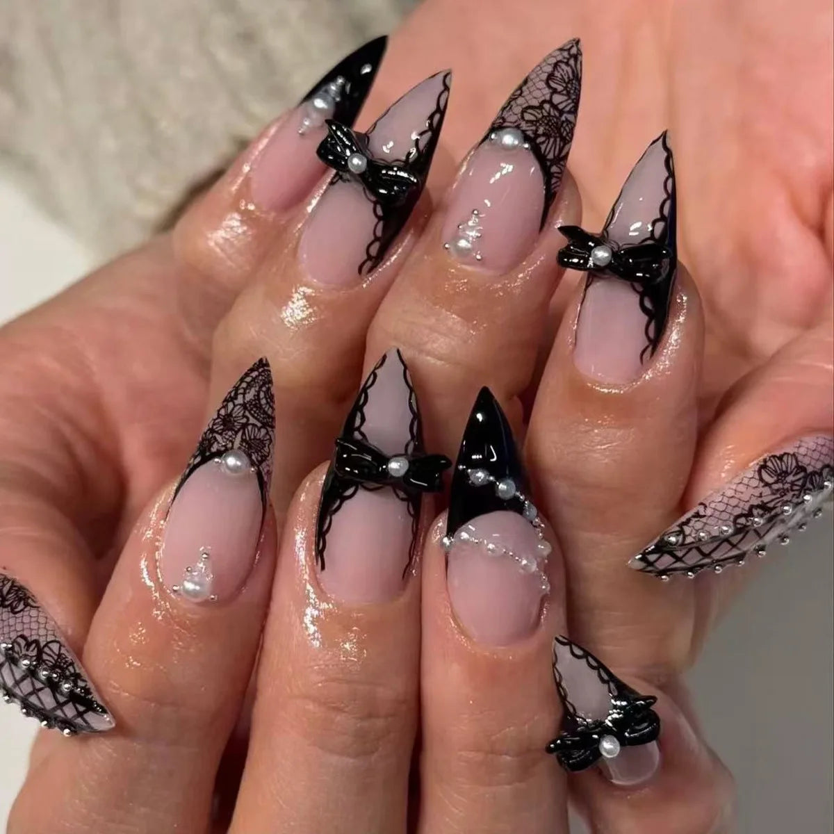Black lace press-on nails worn over pink-nude bases, with pointed dark French tips, raised black bows and small pearl-like accents.