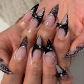 Black lace press-on nails worn over pink-nude bases, with pointed dark French tips, raised black bows and small pearl-like accents.