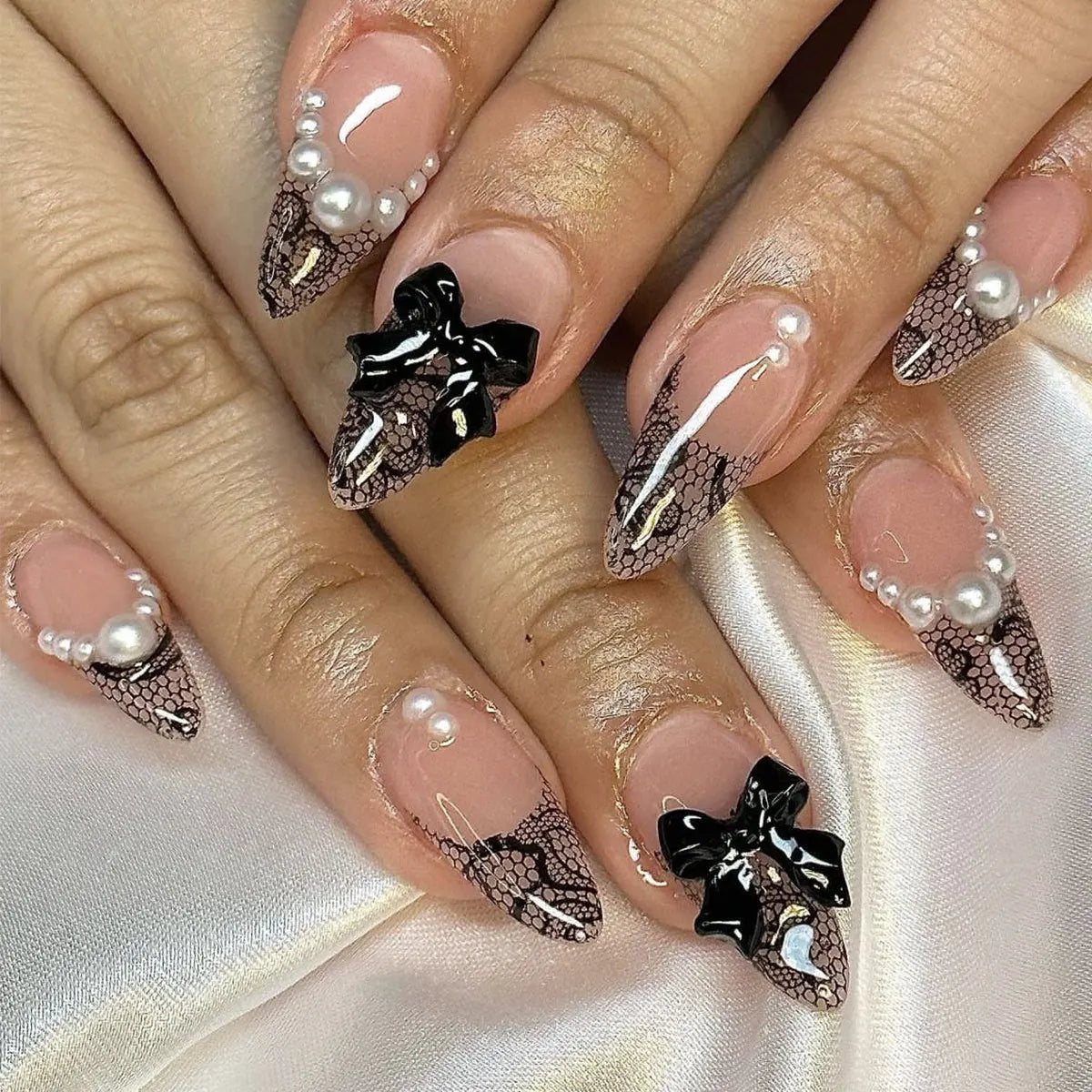Lace French press-on nails displayed on hands over pale satin, with large glossy black bows and clusters of pearl-like accents.