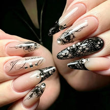Long almond black lace press-on nails worn on both hands, combining nude bases, dark French tips, fine script and small reflective accents against a dark background.