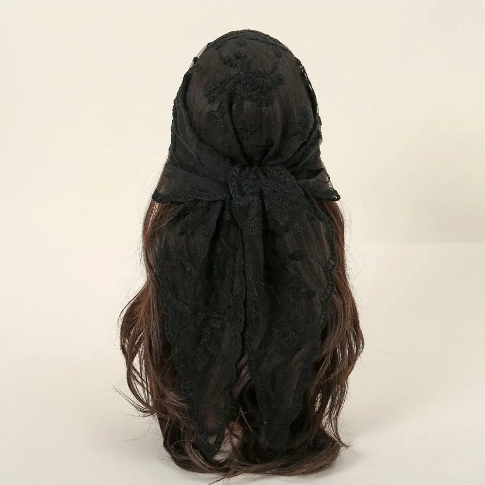 Rear view of a black lace headscarf tied over dark brown hair against a pale background. The knot sits across the back of the head, with the lace ends falling over the hair.