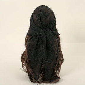 Rear view of a black lace headscarf tied over dark brown hair against a pale background. The knot sits across the back of the head, with the lace ends falling over the hair.