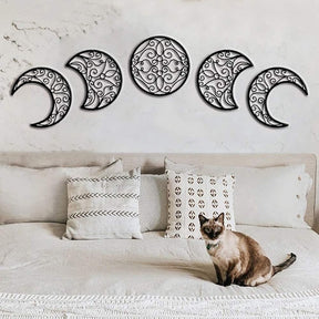 Black openwork moon-phase pieces displayed above a bed with patterned cushions and a cat resting on the bedding.