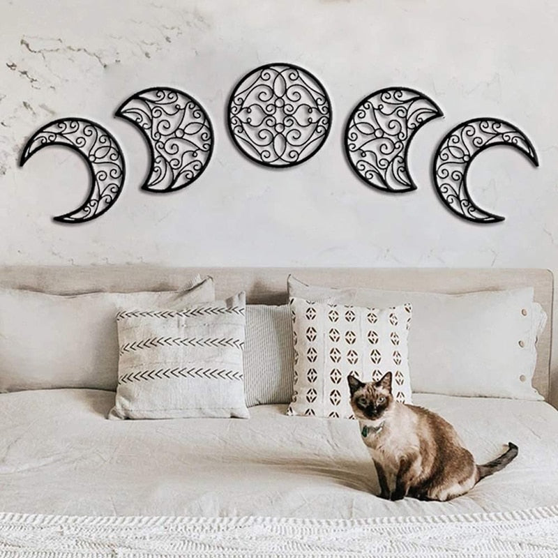 Black openwork moon-phase pieces displayed above a bed with patterned cushions and a cat resting on the bedding.