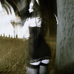 Rear outdoor view of the black dress beside a tree, showing slender shoulder straps, the plain back and lace hem.