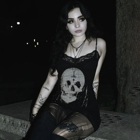 Black skull print lace mini dress worn by a seated model at night. The pale skull, delicate neckline and patterned tights stand out against the dark background.