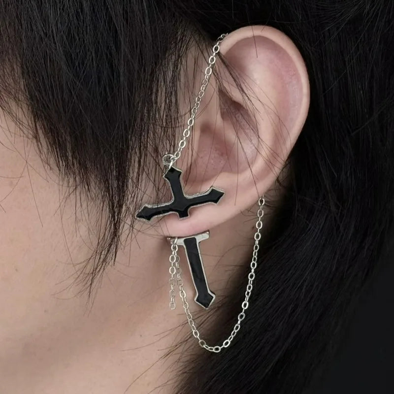 Close view of a black split cross chain earring against dark hair, showing the upper cross above the lobe and the pointed section below it.