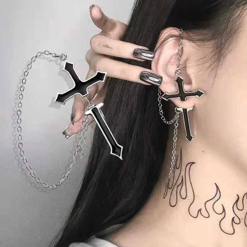 Black split cross chain earring worn on an ear while a second earring is held beside it. Silver-tone edges and looping chains contrast with black hair and metallic nails.