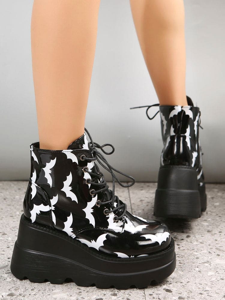 Black platform ankle boots covered in white bat silhouettes, worn with one foot forward to show the glossy print and thick ridged soles.