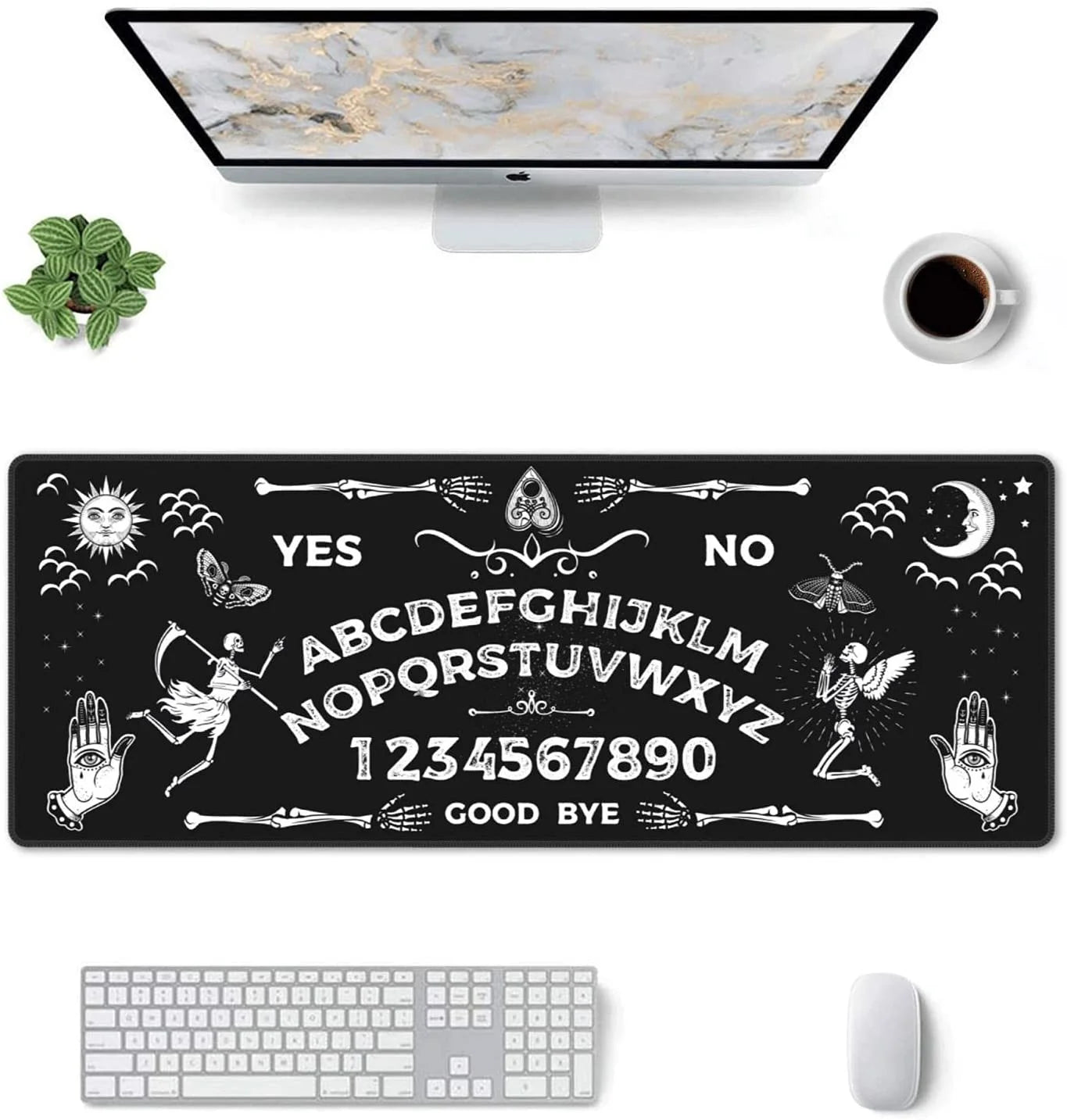 Overhead view of a black-and-white spirit board desk mat arranged with a desktop monitor, keyboard, mouse, plant and coffee cup. Sun, moon and skeleton artwork surrounds the lettering.