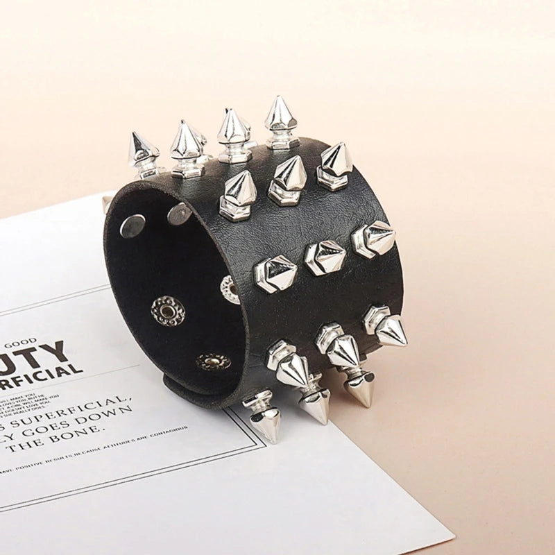 Wide black cuff bracelet angled on a pale surface, showing tall silver-tone spikes, smaller faceted studs and snap fastenings inside the band.
