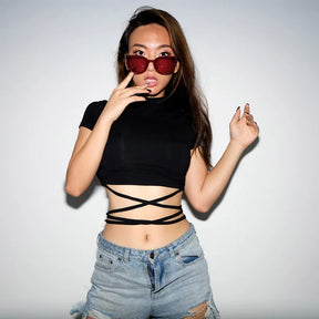 Black waist-wrap crop top worn with denim shorts, with the model touching red sunglasses against a pale studio background.
