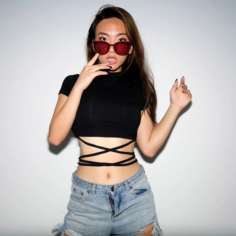Black waist-wrap crop top worn with denim shorts, with the model touching red sunglasses against a pale studio background.