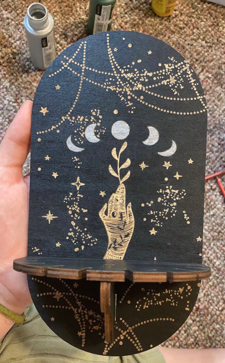 Full view of the compact Moon Phases shelf held upright, showing the rounded black panel, illustrated hand and slotted wooden ledge.