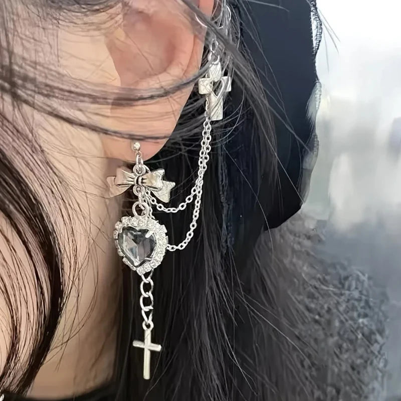 Side view of the clear heart and bow earring beneath a silver-tone upper-ear cuff. Two fine chains connect the pieces and a narrow cross hangs below the heart.