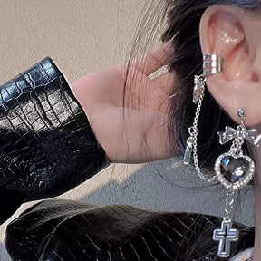 Clear heart chain earring worn beside dark hair with a bow above the sparkling heart and a long cross below. A silver-tone cuff sits at the upper ear beside a glossy black sleeve.
