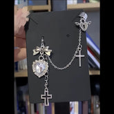 Clear faceted heart and bow earring displayed on a black card. A fine chain connects it to a winged heart cuff with a keyhole shape, and two cross charms hang below.