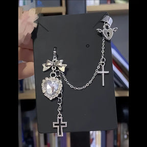 Clear faceted heart and bow earring displayed on a black card. A fine chain connects it to a winged heart cuff with a keyhole shape, and two cross charms hang below.