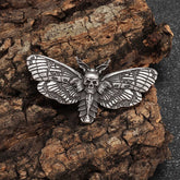 Antique silver tone moth brooch on rough bark, with a small skull at the centre and dark patterned wing recesses.