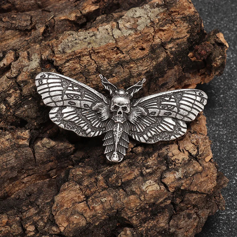 Antique silver tone moth brooch on rough bark, with a small skull at the centre and dark patterned wing recesses.